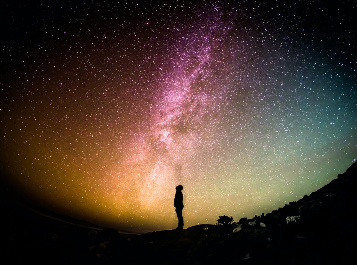 A man stands beneath a vibrant sky filled with stars, creating a stunning contrast against the colorful backdrop