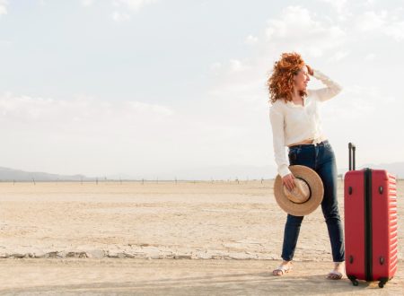 A woman stands in the desert with a suitcase, surrounded by vast sand dunes under a clear blue sky