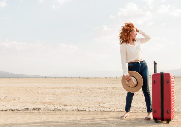 A woman stands in the desert with a suitcase, surrounded by vast sand dunes under a clear blue sky