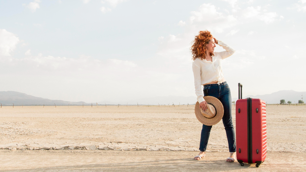 A woman stands in the desert with a suitcase, surrounded by vast sand dunes under a clear blue sky